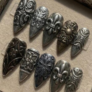 May include: A set of ten press-on nails featuring an antique silver finish with intricate embossed patterns, including fleur-de-lis motifs, floral designs, and scrollwork. Each nail has a pointed almond shape with a metallic, weathered texture.