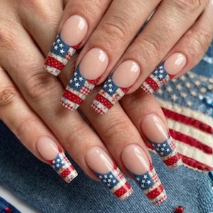 Op de afbeelding: Lange, vierkante nagels met een French tip-ontwerp. De uiteinden hebben een patriottisch patroon van rode en witte strepen en blauw met witte sterren. De nagels zijn geplaatst tegen een denim achtergrond.
