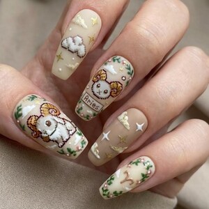 Puede incluir: Una mano con u&ntilde;as largas en forma de almendra con intrincado arte de u&ntilde;as. Los dise&ntilde;os incluyen un carnero pixelado, nubes, estrellas y detalles florales en beige, dorado y verde. Una u&ntilde;a muestra la palabra "Aries".