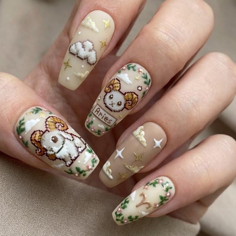 Puede incluir: Una mano con u&ntilde;as largas en forma de almendra con intrincado arte de u&ntilde;as. Los dise&ntilde;os incluyen un carnero pixelado, nubes, estrellas y detalles florales en beige, dorado y verde. Una u&ntilde;a muestra la palabra "Aries".