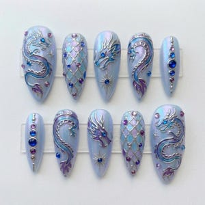 May include: A set of ten light blue, iridescent press-on nails. Each nail features a dragon design in silver, blue, and purple, with jewel accents. The dragon designs vary across the nails, with some featuring a full dragon and others a dragon head.