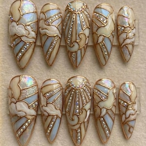 Könnte beinhalten: Ein Set aus zehn mandelförmigen Kunstnägeln. Jeder Nagel zeigt ein Buntglasdesign mit goldenen Konturen, schillernden Akzenten und eingebetteten Strasssteinen. Das Design beinhaltet wolkenartige Formen und Sonnenstrahlenmuster in einer hellblauen und cremefarbenen Farbpalette.