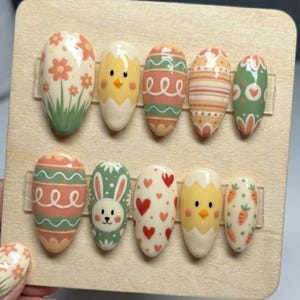 Può includere: Un set di dieci unghie finte a tema pasquale. I disegni includono fiori, pulcini, uova con strisce e linee ondulate, un coniglietto e cuori. Le unghie ovali sono dipinte in colori pastello, su un espositore in legno.