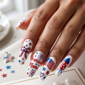 May include: A hand with long, white, decorated fingernails. Each nail features a different patriotic design in red, white, and blue, including a bow, stars, fireworks, an American flag, and the words "American Mama."