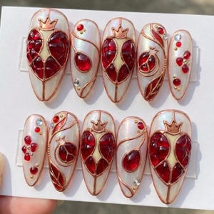 Puede incluir: Un conjunto de diez uñas postizas en forma de almendra. Cada uña presenta un diseño de granada con una corona, gemas rojas y detalles dorados. Las uñas tienen una base blanca nacarada.