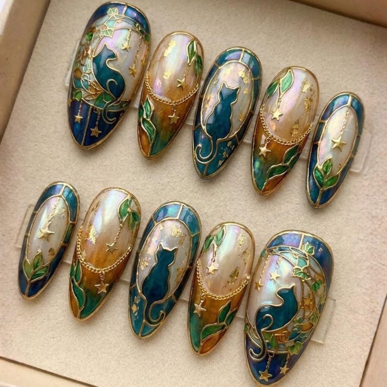 May include: A set of ten oval-shaped, painted fingernails. Each nail features a teal cat silhouette, gold stars, and gold chain accents. The nails have a pearlescent base with teal, gold, and green details, and a gold border.