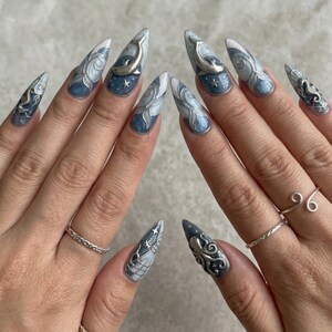 K&ouml;nnte beinhalten: Lange, spitze Fingern&auml;gel mit einem blau-wei&szlig;-silbernen Ozean-Design. Die N&auml;gel zeigen Wellenmuster, Sterne und silberne Delfin- und Seepferdchen-Verzierungen. Die N&auml;gel sind mit silbernen Ringen verziert.