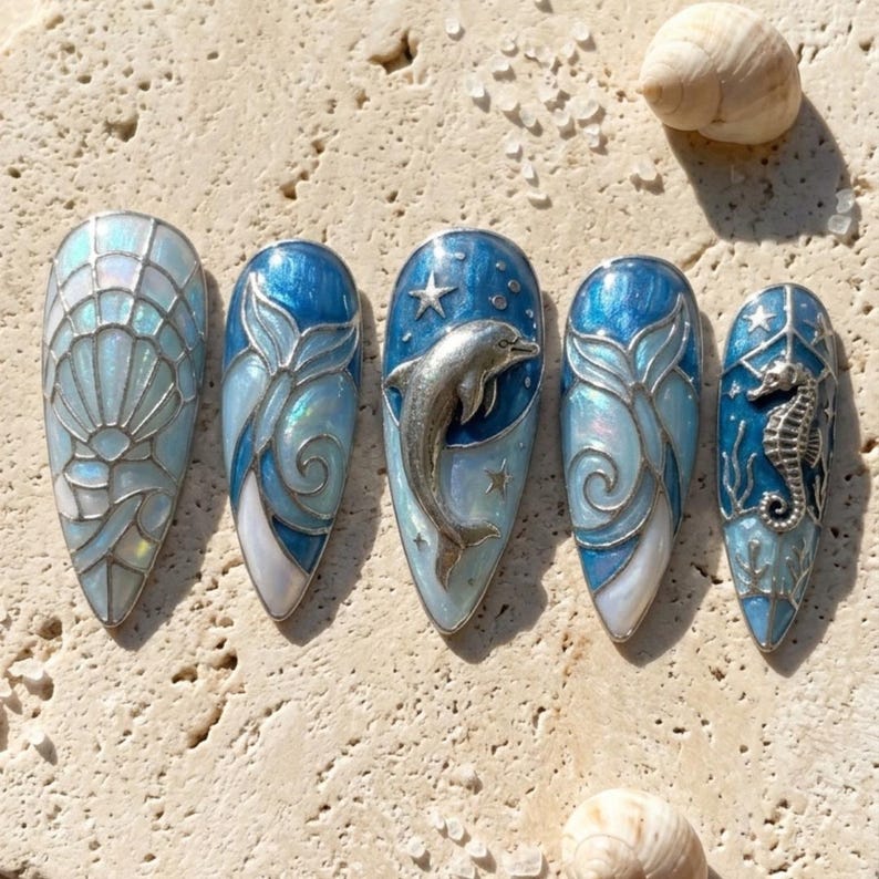 K&ouml;nnte beinhalten: Ein Set aus f&uuml;nf langen, mandelf&ouml;rmigen Kunstn&auml;geln mit Meeresmotiven. Jeder Nagel zeigt ein anderes Design, darunter eine Muschel, ein Delfin und ein Seepferdchen, in Blau-, Silber- und Wei&szlig;t&ouml;nen. Die N&auml;gel haben ein gl&auml;nzendes Finish und sind mit aufwendigen Details verziert.