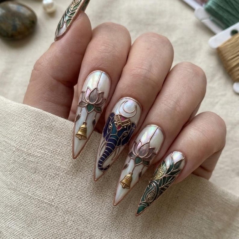 May include: Long, pointed fingernails with intricate designs. The nails feature a pearlescent base with detailed illustrations of a lotus flower, an elephant, and ornate patterns. Gold bells and accents add to the decorative style.