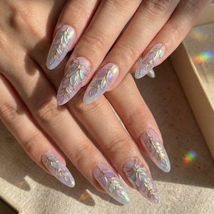May include: Long, almond-shaped nails with a pearlescent finish. Each nail features a gold-colored leaf design with iridescent accents of purple, blue, and green. The nails are well-manicured and appear to be professionally done.
