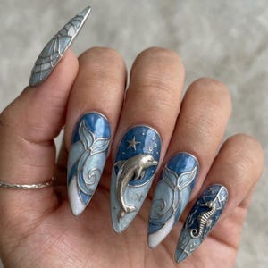 K&ouml;nnte beinhalten: Lange, mandelf&ouml;rmige N&auml;gel mit maritimem Thema. Die N&auml;gel zeigen einen Farbverlauf von Blau und Wei&szlig; mit silbernen Delfin-, Seepferdchen- und Schwanzmotiven. Ein Nagel hat ein silbernes Seepferdchen, ein anderer einen silbernen Delfin.
