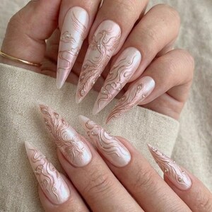 May include: Long, pointed, iridescent nails with a pearlescent finish. Each nail features a detailed design in a rose gold hue, including phoenixes, swirling patterns, and small pearl accents. The nails are a light pink color.