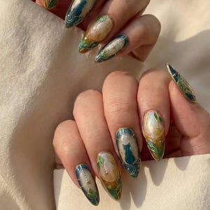 May include: Almond-shaped nails with intricate designs. Each nail features a unique artwork, including a blue cat, stars, and botanical elements. The color palette includes teal, gold, and iridescent pearl, creating a sophisticated and artistic look.
