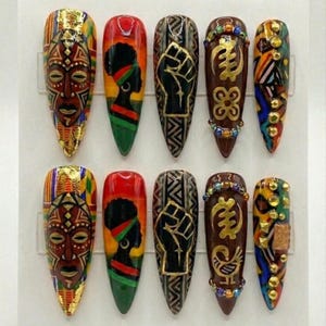 Pode incluir: Um conjunto de dez unhas acrílicas longas e pontiagudas com vários designs de inspiração africana. Os designs incluem máscaras tribais, silhuetas de mulheres, punhos erguidos e símbolos Adinkra, em ouro, vermelho, verde e preto.