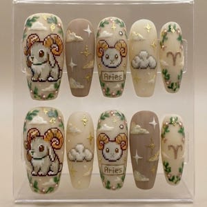 Puede incluir: Un conjunto de diez u&ntilde;as artificiales con tem&aacute;tica del signo zodiacal Aries. Las u&ntilde;as presentan ilustraciones pixeladas de carneros, nubes, estrellas y la palabra "Aries". Los colores incluyen beige, marr&oacute;n, dorado y verde, con detalles florales y de hojas. Se exhiben en una caja de pl&aacute;stico transparente.