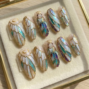 May include: A set of ten artificial nails with an angel wing design. The nails feature a pearlescent base with gold accents and iridescent teal, purple, and white details. Some nails have a small angel figure with a halo.