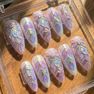 May include: A set of ten artificial nails with an oval shape. Each nail is painted with a pearlescent white base and adorned with a gold-colored leaf design, featuring iridescent purple and blue accents. The nails are arranged on a wooden surface.