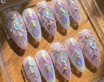 May include: A set of ten artificial nails with an oval shape. Each nail is painted with a pearlescent white base and adorned with a gold-colored leaf design, featuring iridescent purple and blue accents. The nails are arranged on a wooden surface.