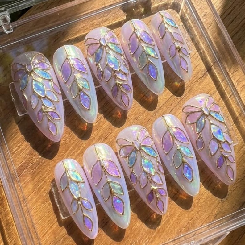 May include: A set of ten artificial nails with an oval shape. Each nail is painted with a pearlescent white base and adorned with a gold-colored leaf design, featuring iridescent purple and blue accents. The nails are arranged on a wooden surface.