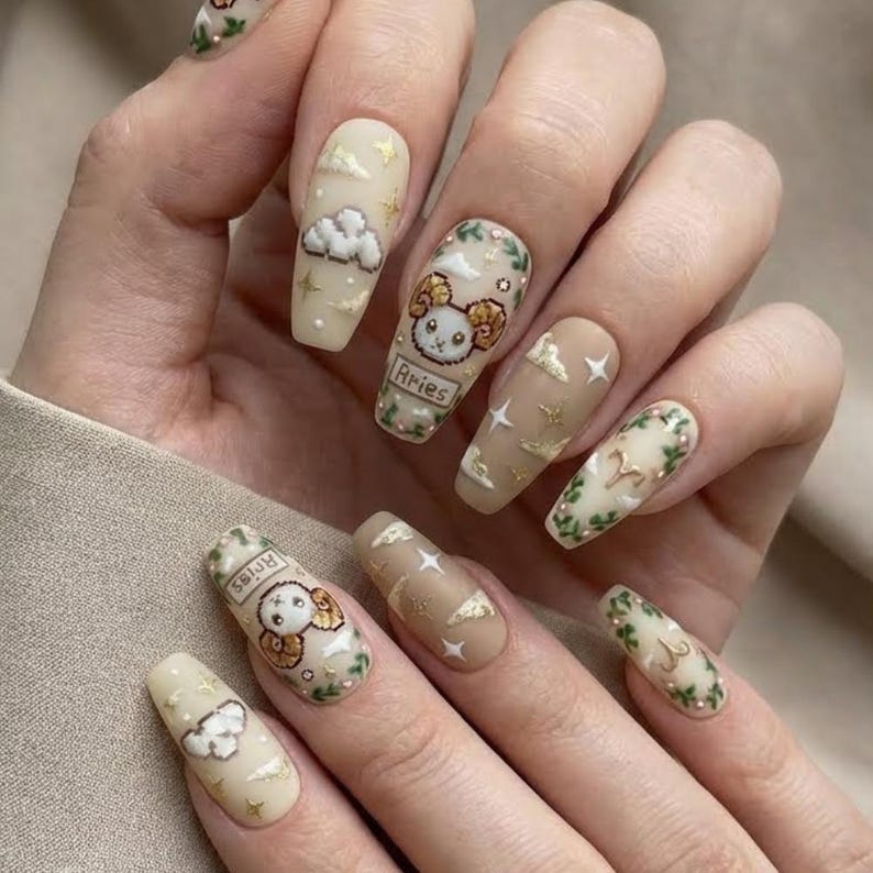 Puede incluir: Una mano con u&ntilde;as largas en forma de almendra. Las u&ntilde;as est&aacute;n pintadas con varios dise&ntilde;os, incluyendo nubes, estrellas y un s&iacute;mbolo del zodiaco Aries. La paleta de colores incluye beige, dorado y verde.