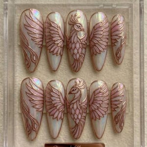 May include: A set of ten almond-shaped press-on nails featuring an iridescent white base with raised rose gold phoenix designs. Each nail displays intricate wing patterns and stylized bird motifs with a metallic finish, arranged in a clear plastic case.