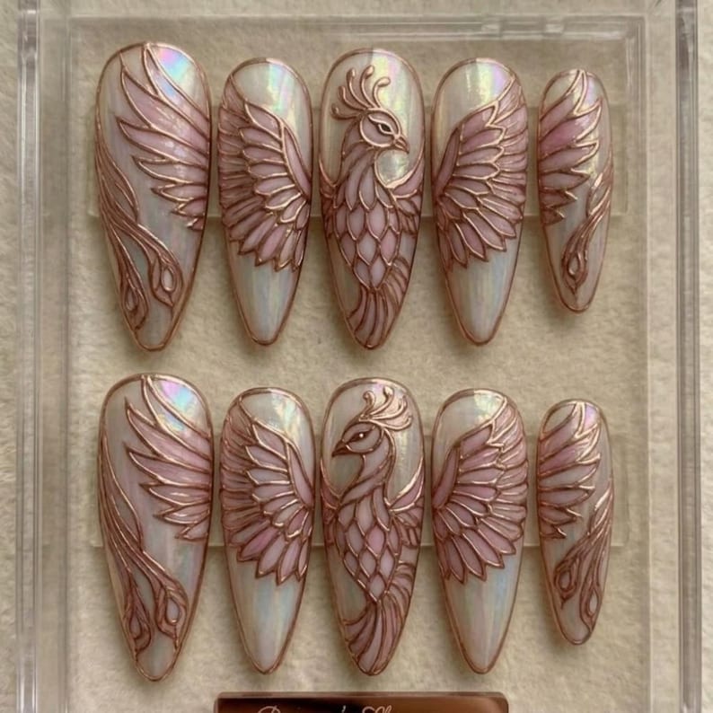 May include: A set of ten almond-shaped press-on nails featuring an iridescent white base with raised rose gold phoenix designs. Each nail displays intricate wing patterns and stylized bird motifs with a metallic finish, arranged in a clear plastic case.