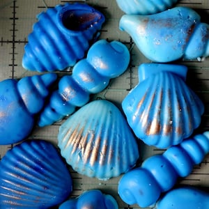 May include: A collection of blue seashell-shaped objects with a metallic gold accent. The shells vary in size and shape, with ribbed and smooth textures. The objects are arranged on a grey surface with grid lines.