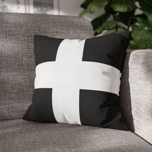 May include: Black and white decorative throw pillow with a bold white cross design. The pillow is square-shaped and rests on a gray sofa. The contrasting colors create a striking visual effect, suitable for home decor.
