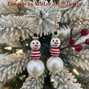 May include: A pair of snowman earrings with gold-tone hooks. Each earring has a white bead head with an orange nose and black hat, a red and white striped bead body, and a large white pearl bead base. The text "Earrings by MDLee Art & Design" is visible.