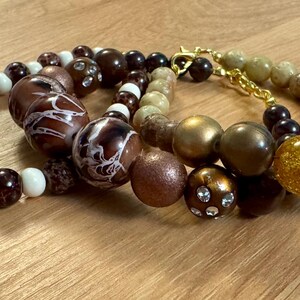 Pode incluir: Uma coleção de pulseiras de contas em tons de castanho, dourado e branco. As pulseiras apresentam vários tamanhos e texturas de contas, incluindo algumas com glitter e detalhes de strass. Uma fivela de corrente dourada é visível.
