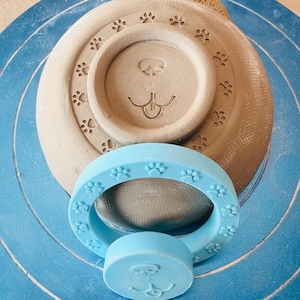 May include: A light brown clay bowl featuring an embossed animal nose and mouth design at the centre, surrounded by a ring of paw prints. A light blue plastic stamp tool with a matching paw print border and nose design sits in front.