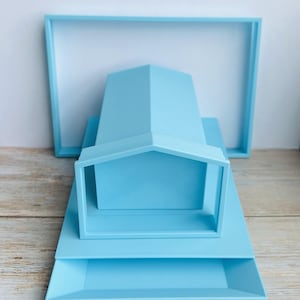 May include: A set of light blue geometric shapes, including a house-shaped frame, a rectangular tray, and a square frame. The objects are arranged on a wooden surface against a white background. The shapes are all the same shade of blue.