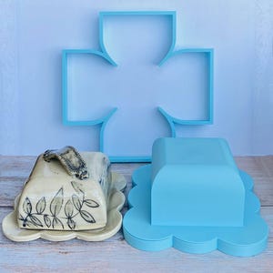 Peut inclure: Un beurrier en céramique avec un vernis crème et des illustrations de feuilles noires, doté d'une poignée sculptée. À côté se trouve un couvercle de beurrier en plastique bleu clair uni et un cadre d'étagère mural en forme de croix.