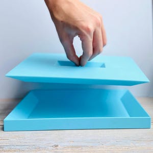 Peut inclure: Un récipient carré bleu clair avec un couvercle assorti. Le couvercle comporte une découpe de poignée rectangulaire. Une main soulève le couvercle, révélant l'intérieur vide du récipient. Le récipient repose sur une surface en bois de couleur claire.