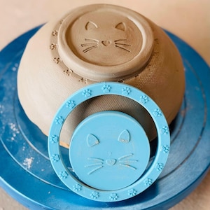 Peut inclure: Un tampon à poterie circulaire bleu clair présentant un motif de visage de chat central entouré d'un anneau de petites empreintes de pattes, placé devant un bol en argile beige inversé affichant un motif identique de chat et de pattes.
