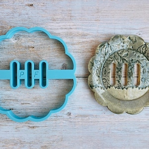 May include: A light blue plastic cookie cutter in a scalloped shape with the letters "PF" and three vertical bars. Beside it is a floral-patterned soap dish with three slots.