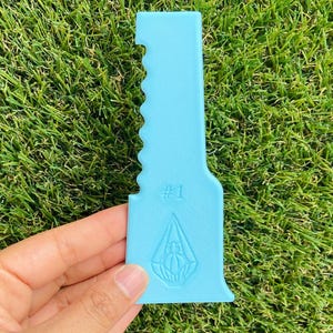 May include: A light blue plastic tool with a serrated edge on one side, featuring the text #1 and a diamond-shaped emblem with a spider graphic embossed on the lower section. The tool has a flat, rectangular top and a wider, curved base.