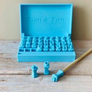 May include: A bright blue plastic storage case with the text Pearl & Fern embossed on the lid, containing a set of individual letter stamps. Two loose letter stamps and a thin wooden handle with a blue stamp attachment are placed in front.