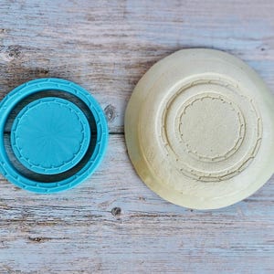 May include: A bright turquoise circular plastic cookie cutter with a scalloped edge and a matching circular insert, positioned next to a round, off-white textured ceramic dish featuring a concentric circular pattern on its base.