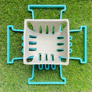 May include: A square, light grey planter with rectangular cut-outs is surrounded by a turquoise geometric support structure. The planter and support are set against a backdrop of green grass.