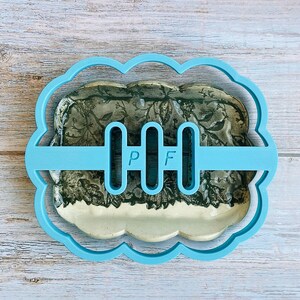 May include: A light blue cookie cutter with a scalloped edge, holding a floral-patterned cookie. The cutter has three vertical bars with the letters "P" and "F" on the center bar. The cookie has a white base.