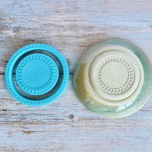 May include: A bright turquoise plastic circular mould with a textured rim featuring a repeating teardrop pattern, positioned next to a round ceramic bowl with a matching teardrop-patterned base in a light beige and sage green glaze.