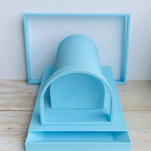 Puede incluir: Un conjunto de exhibición minimalista azul claro que presenta un marco rectangular, una estructura arqueada y una bandeja rectangular. Los objetos están dispuestos sobre una superficie de vetas de madera clara, creando una estética limpia y moderna.