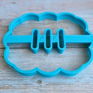 May include: A bright turquoise cookie cutter shaped like a rugby ball, featuring three vertical bars with the letters "P" and "F". The cutter has a scalloped edge. The item is likely used for baking.