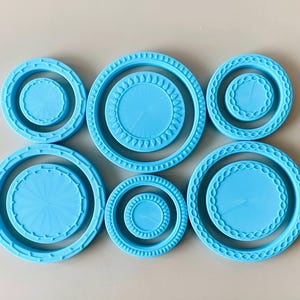 May include: A set of six light blue circular silicone moulds arranged in two rows. Each mould features a distinct textured border pattern, including radial lines, small teardrop shapes, and interlocking chain-link designs on the inner rims.