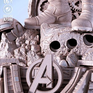 May include: A detailed, light gray statue featuring the Avengers logo, boots, and a helmet. The statue is made of resin and FDM, with a scale of 1/6. The design includes a rocky, textured base and architectural elements.