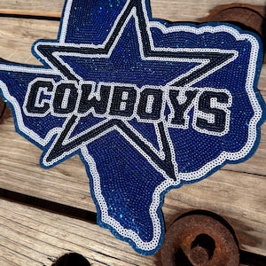 May include: A sequined patch shaped like the state of Texas featuring a large blue star outlined in white and dark blue. The word COWBOYS is written in bold white letters with a dark blue outline across the center of the star and state shape.
