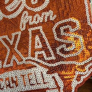 May include: A decorative sign shaped like the state of Texas, covered in shimmering orange sequins. The text 'Greetings from TEXAS' is embroidered in white sequins across the surface, with the word 'TEXAS' prominently displayed in large capital letters.