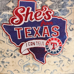 May include: A Texas-shaped sequin patch featuring the red text She's from TEXAS and I CAN TELL on a blue background. A circular Texas Rangers baseball logo with a red T and baseball stitching is positioned on the lower right side of the patch.