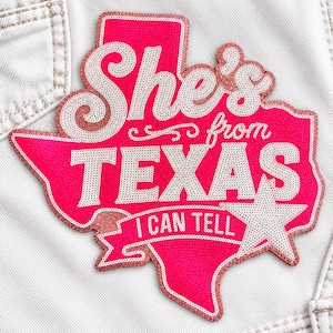 May include: A Texas-shaped patch featuring bright pink sequins as the base color. The text "She's from TEXAS I CAN TELL" is embroidered in white sequins across the center. A white sequined star is positioned on the lower right side of the patch.
